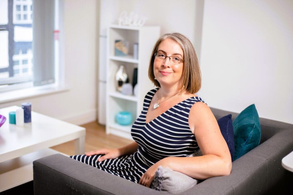 Hannah Paskin in her counselling room in Cheshire. She is posed for a photographer sat on a sofa. She is turned back towards the camera and smiling. In the background you can see her counselling room with a table and bookcase.
