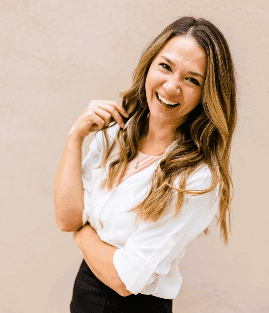 A woman standing. She is laughing looking at the camera. She has long dark blonde hair which is styled curly. She has her arms crossed. She wears a white blouse and block bottoms.
