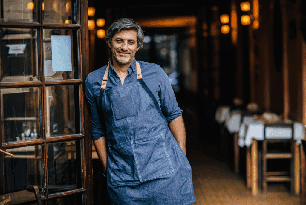 A man stands in a doorway. He wears an apron suggesting his work is something where he makes things, the building behind is perhaps a cafe or bakery. He has grey hair and wears a blue shirt under his apron. There is a wooden door next to him
