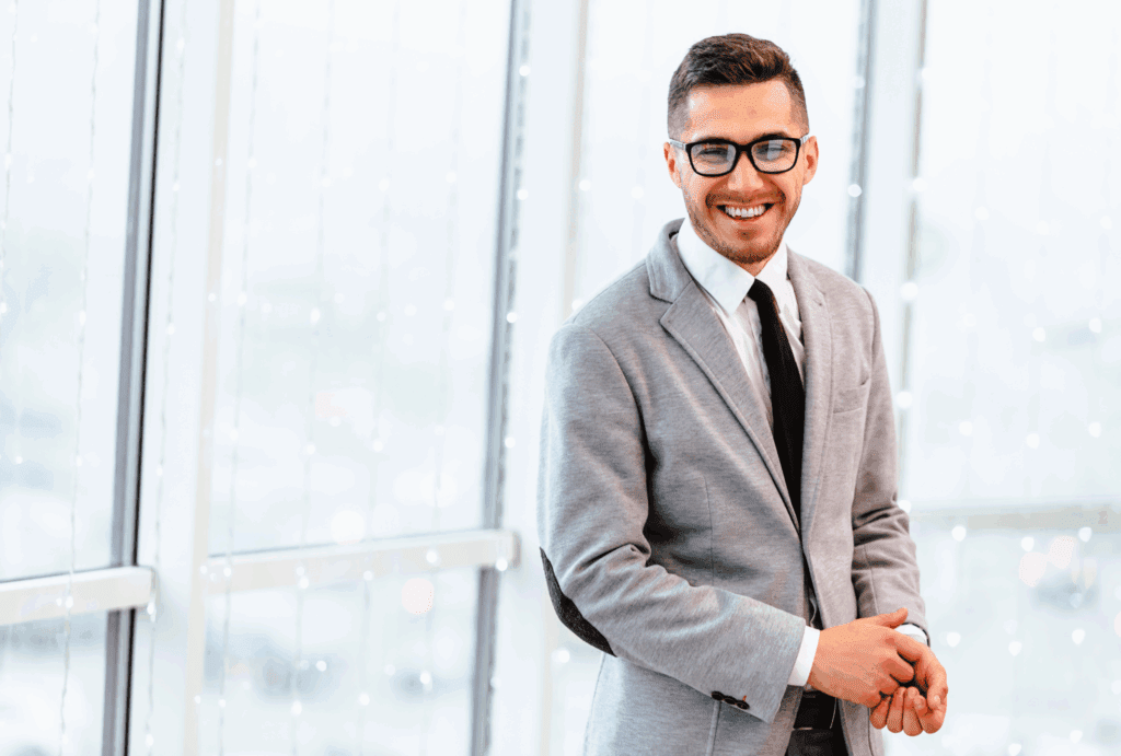 Male stood in front of glass windows of an office building. He has dark hair and wears glasses. He has a white shirt, dark tie and grey jacket. He is looking into the distance and smiling