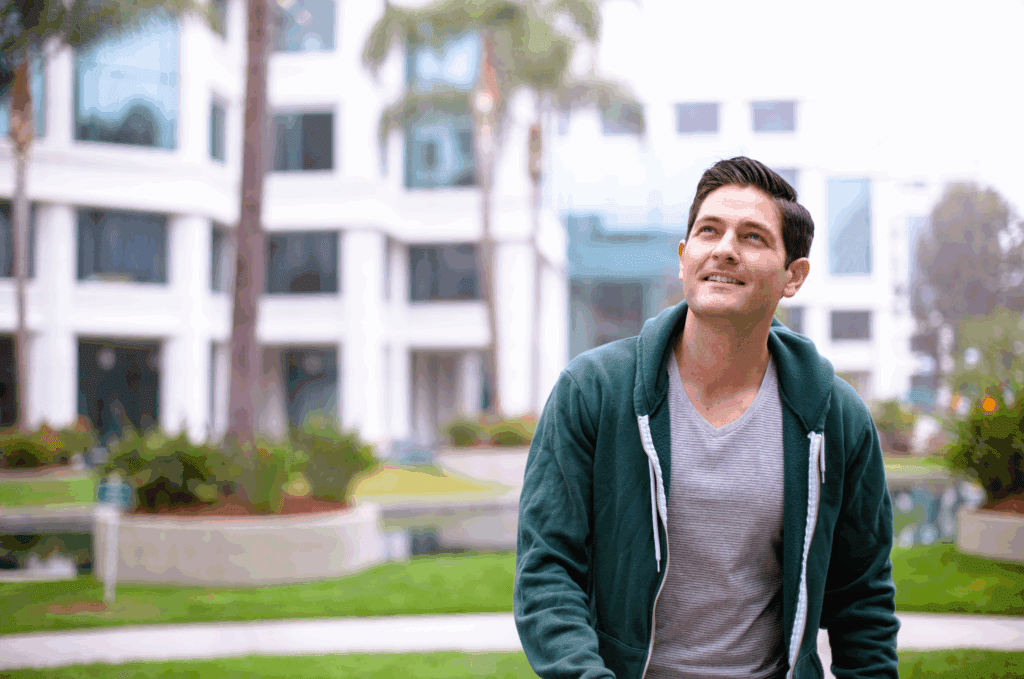 Male sat outside, likely on a bench. There are out of focus buildings in the background. He wears a green zip up hoodie. He has dark hair. he is smiling and looking into the distance
