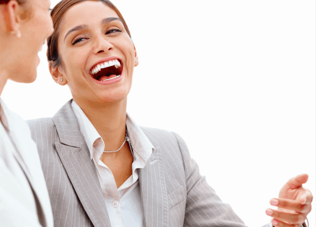 2 females in the image, both laughing with each other. They are both dressed in work suit attire and both have their hair tied back.