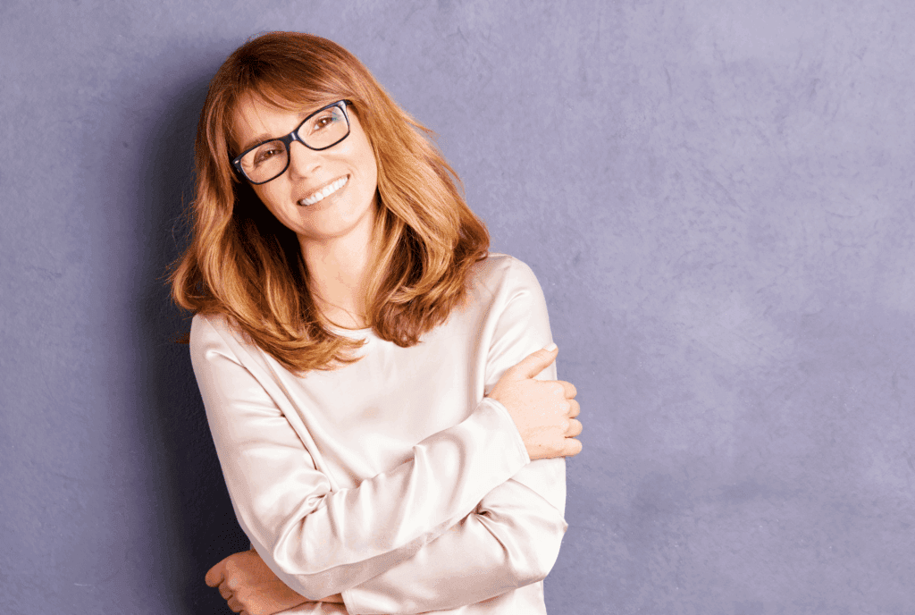 A female leaning against a wall. The wall is pale blue. She wears a cream long sleeved casual top. She has long thick dark blonde hair and wears glasses. She stands with arms crossed